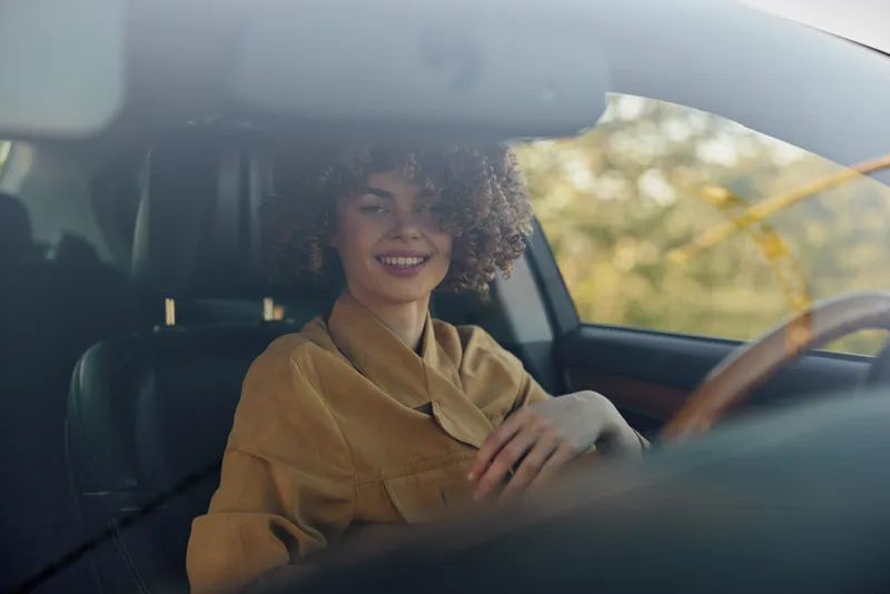 A young woman with curly hair sits in a car, smiling softly as autumn light floods the seat, capturing a candid, relaxed moment with a retro film color mood that evokes nostalgia today.