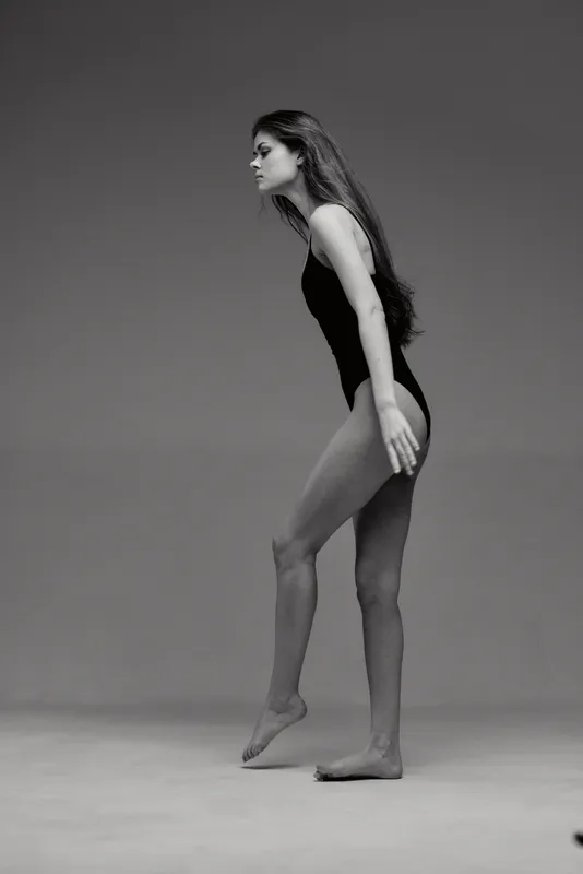 Black and white image of a slender woman balancing on one foot in a studio setting, expressing poise and grace with long hair and minimal clothing.