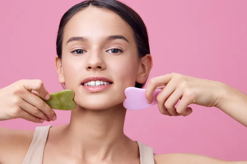 Young woman using facial massage tools on her cheeks, promoting healthy skin and beauty trends Soft pink background enhances a fresh and joyful atmosphere