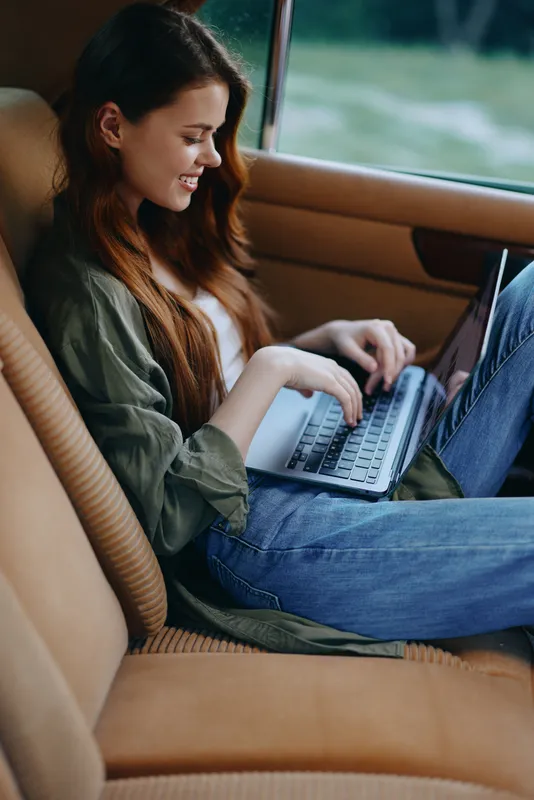 Young woman with long red hair working on a laptop in a car, wearing casual clothing, with a joyful expression, in a natural outdoor setting.