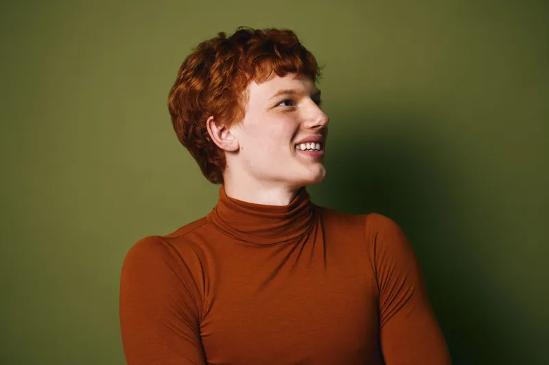 Portrait of young redhead man with curly hair smiling wearing brown turtleneck sweater against green background. Casual style and happy emotion in studio lighting.