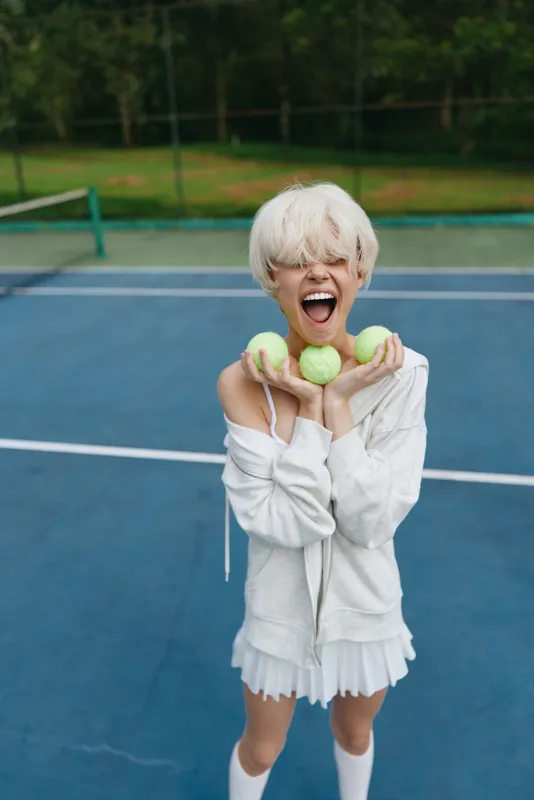 Happy blonde woman with a playful expression holding green tennis balls on a tennis court in summer, wearing a stylish white sports outfit
