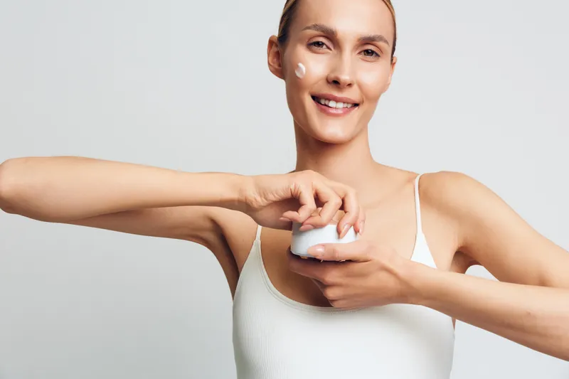 Smiling young woman in a white tank top applies face cream with her fingers against a plain light background, showing a skincare routine and natural beauty concept.