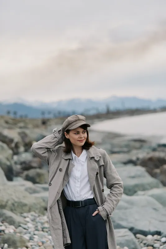 Young woman in checkered coat and cap stands on rocky beach with distant mountains and cloudy sky. Casual fashion outdoors, autumn style and natural landscape concept.