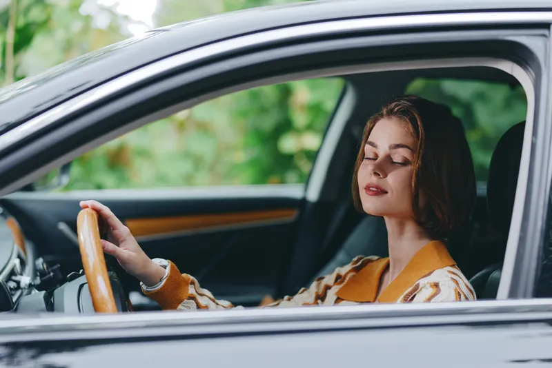 A woman sits behind the wheel in a calm moment, smiling softly as she grips the steering wheel with confidence, ready for a journey, enjoying the quiet focus of driving in a stylish car interior.