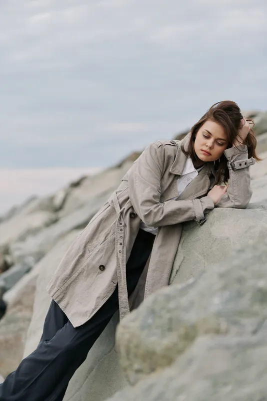 Young woman in beige trench coat leans on large rock outdoors, eyes closed, enjoying calm moment in natural rocky landscape. Concept of relaxation, fashion, and nature connection.