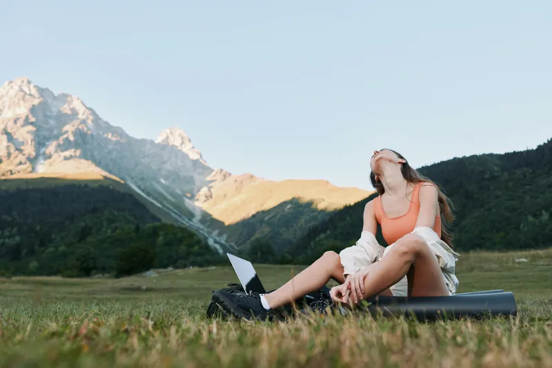 A relaxed young woman sits on a grassy field with a laptop, enjoying nature under a clear sky. She wears a sleeveless top and smiles, exuding calm focus as she works amidst the open countryside.