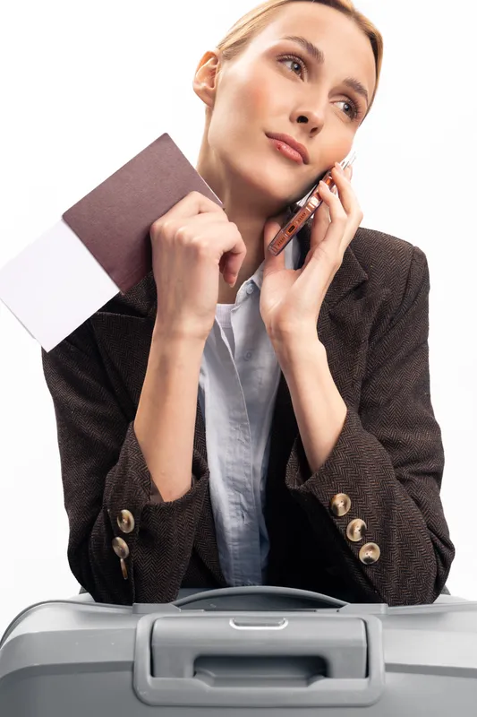 Young woman in blazer holds passport and talks on phone while waiting with suitcase. Travel concept with businesswoman planning trip and looking thoughtfully isolated on white background.