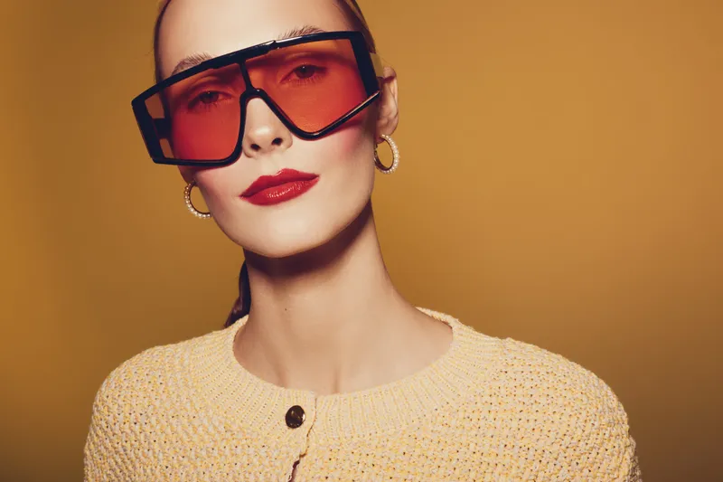 Stylish woman with red lipstick and large orange sunglasses poses against yellow background wearing beige knitted sweater and gold hoop earrings. Fashion portrait with confident mood.
