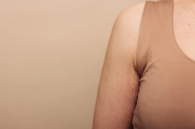 Close-up of a woman's upper arm and shoulder in a beige sleeveless top against a plain beige background. Concept of natural skin texture, body positivity, and minimalistic fashion.