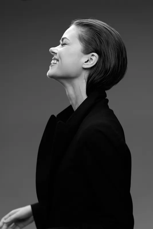 Smiling woman in black coat with elegant hairstyle, black and white portrait, joyful expression, studio background, monochrome fashion.
