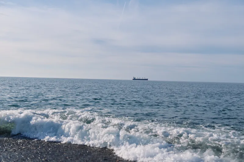Sea view with distant ship on the horizon, gentle waves rolling to a pebbled shore, clear sky above, a tranquil maritime scene ideal for travel, nature, and exploration themes.