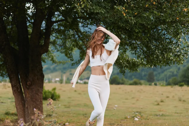 Cheerful woman outdoors in a white outfit, hair loose, sunlit meadow moment, relaxed pose, gentle breeze, natural colors, vibrant greenery background