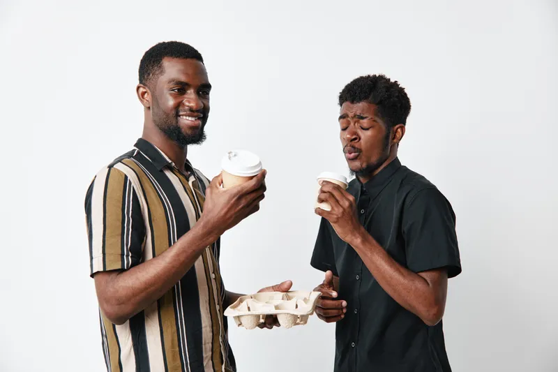 Two African American men with dark skin holding coffee cups and a drink carrier, smiling and enjoying beverages in casual clothing against white background.