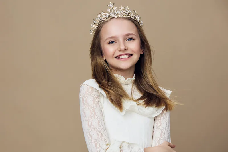 A joyful young girl in a white lace dress and a crown, smiling confidently against a warm beige background, perfect for children s photography and portraits