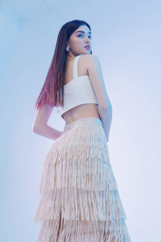 Young woman in a white crop top and layered fringe skirt poses confidently against a soft blue background. Concept of woman beauty fashion with modern style and elegance.