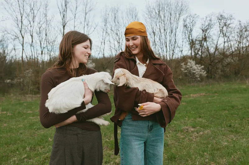 Two women in a rural field share smiles while holding young goats, capturing farm life and companionship. Natural daylight highlights playful interaction, simple clothing, and a wholesome, relaxed