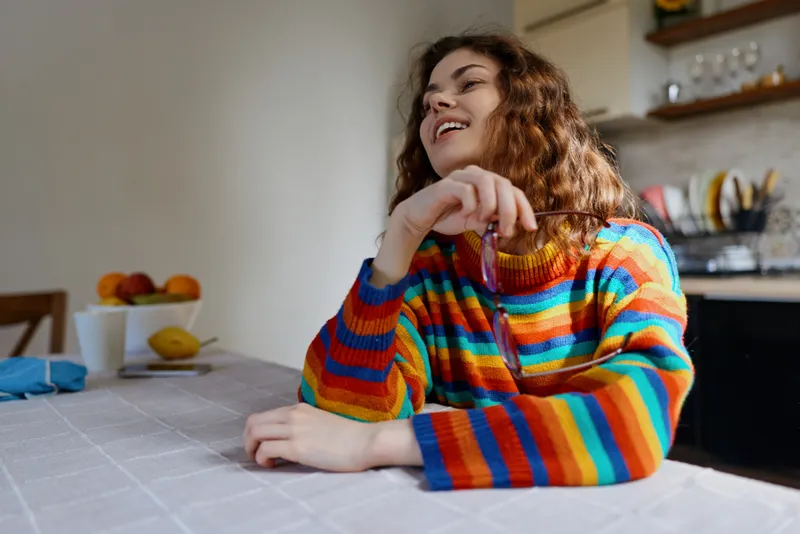 Cheerful woman indoors, seated at a dining table, wearing a rainbow striped sweater, soft natural light, cozy home ambiance, relaxed posture, everyday life vibe, gentle smile, warm colors