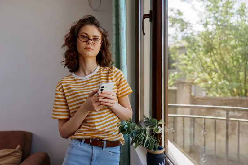 A young woman with curly hair and glasses stands by a sunny window, looking at her smartphone in a casual striped tee and jeans, conveying a relaxed, everyday home lifestyle moment.
