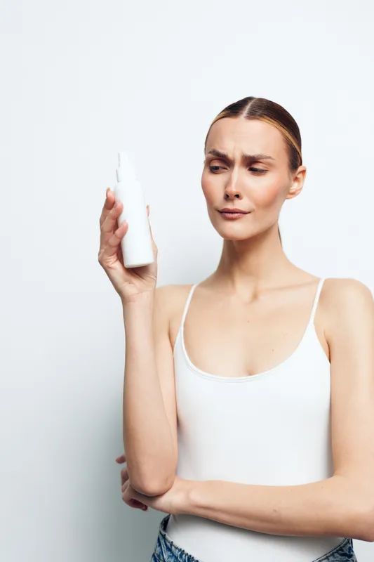 Young woman in white tank top looks skeptically at white spray bottle holding it close to face isolated on plain background with thoughtful expression and crossed arms.