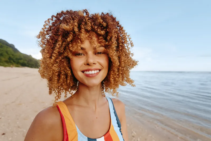 Smiling young woman with curly hair stands on a sunny beach wearing a colorful striped swimsuit, with a calm sea and blue sky behind, conveying confidence and carefree mood.