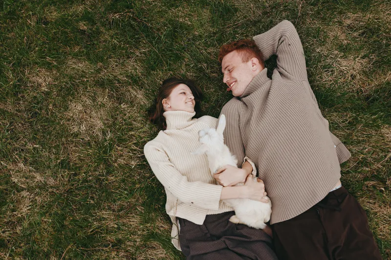 Couple lying on grass with a small white goat kid, cozy sweaters, and a gentle moment outdoors. Lifestyle scene highlighting bonding, family, and nature, with soft sunlight and relaxed, intimate mood.