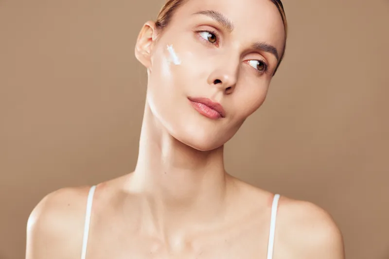 Young woman with clear skin applies moisturizing cream on face looks away with gentle expression isolated on beige background in skincare beauty routine concept.