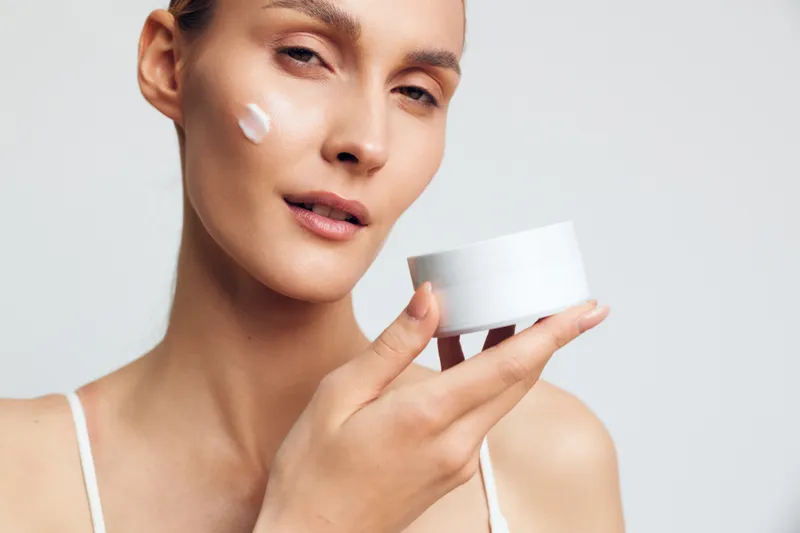 Young woman applying white face cream on cheek holding cosmetic jar. Skincare routine concept with natural makeup on light background. Soft lighting highlights healthy skin texture.