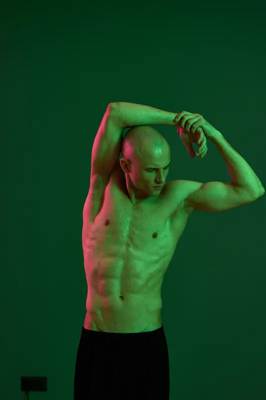 Athletic man stretching in dramatic green and red lighting, showcasing a strong physique and focused expression in a dynamic pose.