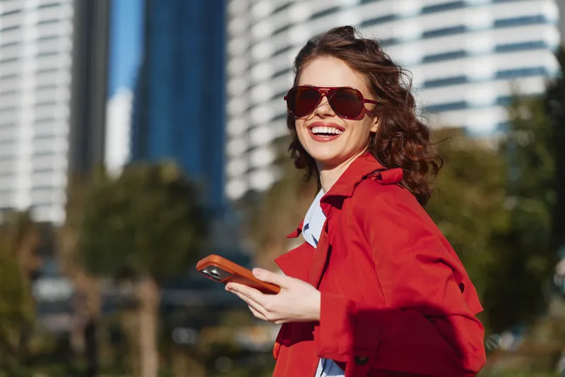 A young woman with curly hair wearing a red jacket and sunglasses smiles while holding a smartphone outdoors in an urban setting on a sunny day.