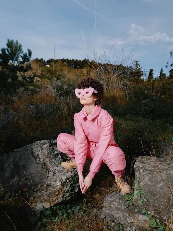 Person in a pink jumpsuit sits on rocky terrain in an outdoor nature setting, floral sunglasses and a dreamy pose, artistic lifestyle photo with natural background.