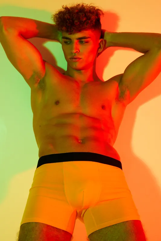 Young man with muscular body posing in colorful lighting wearing yellow boxer briefs, fitness and fashion concept with confident expression and casual style.