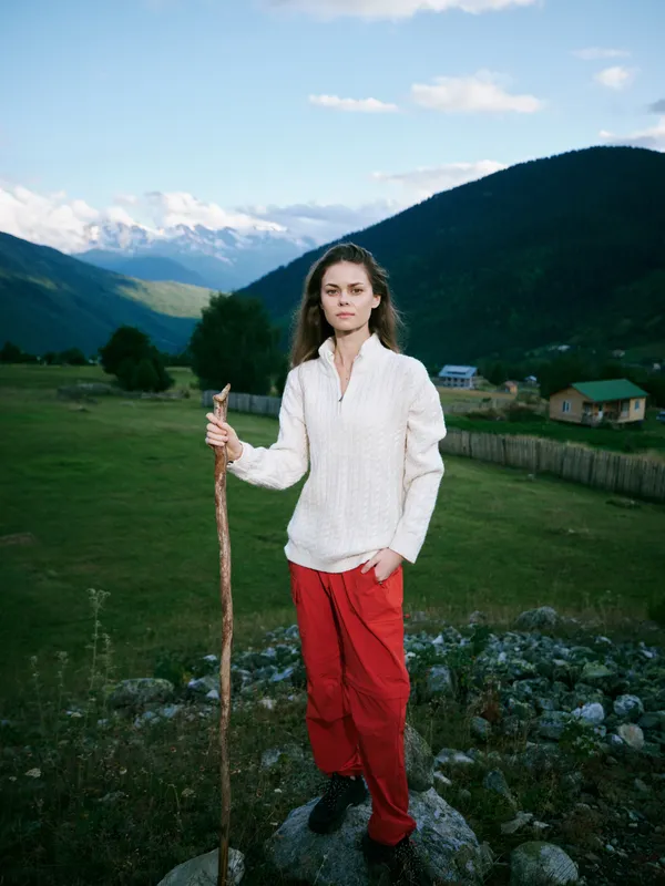 Woman hiking in a mountain countryside with a walking stick, wearing a white sweater and red pants, standing on rocks as alpine scenery and a distant village frame a tranquil outdoor landscape