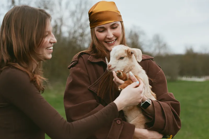 Goat, kid, woman outdoors, smiling, holding a young animal while friend shares the moment, casual jacket, natural light, grassy field, candid lifestyle, warm colors, connection with nature