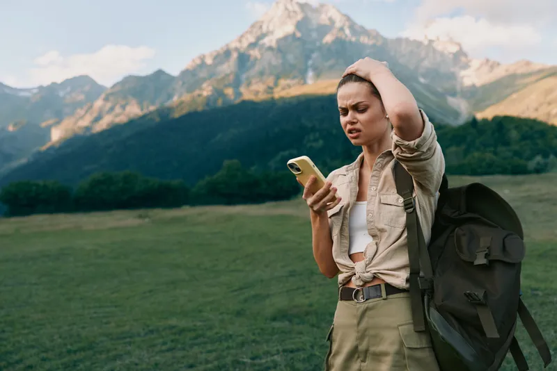 Cheerful woman outdoors, embracing a casual lifestyle with a backpack, checking a smartphone amid mountains and wide green fields, sunlit scene, serene mood in natural tones