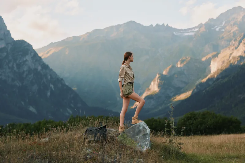 A woman stands on a rock in a wide meadow with distant mountains, embodying outdoor lifestyle and nature, a calm moment of adventure, exploration, and scenic beauty.