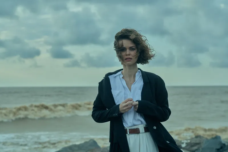 Woman stands on a windy beach in a blazer and white shirt, ocean behind, moody clouds, casual chic style and sea breeze as waves crash nearby