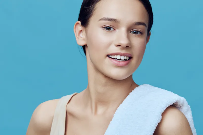 Young female model with a towel on her shoulder, showcasing a fresh and healthy look against a blue background, perfect for wellness and beauty concepts