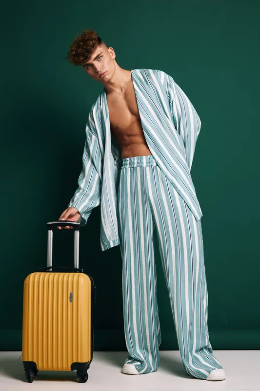 Young man in striped pajamas standing with yellow suitcase on wheels against green background, casual travel style, relaxed pose and modern luggage for trip or vacation.