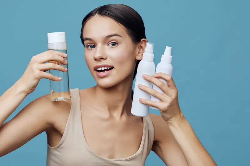 Young woman promoting skincare products, smiling while holding three different bottles against a light blue background Perfect for healthy skin concepts