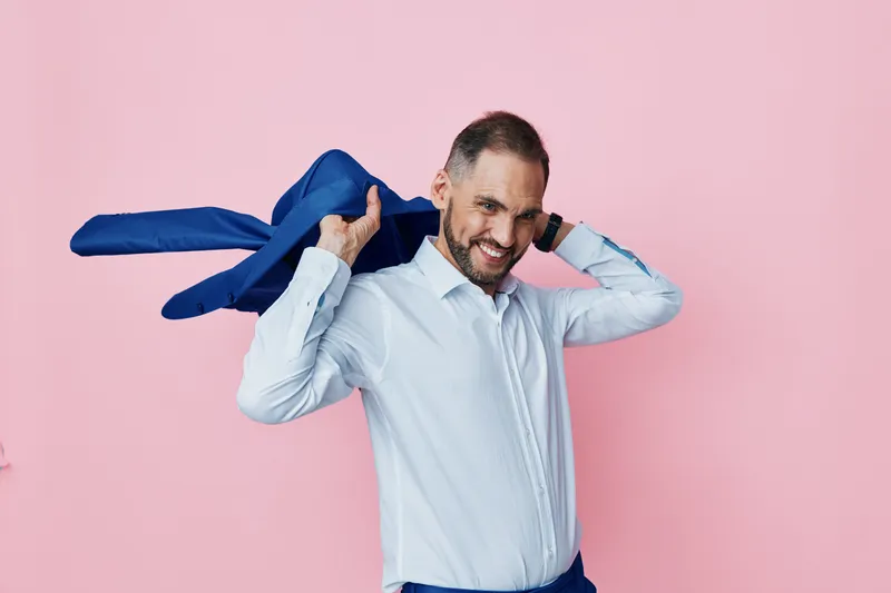 A business professional man stands against a soft pink background, projecting confidence and focus. He slings a jacket over his shoulder, signaling casual executive style and prepared resolve.