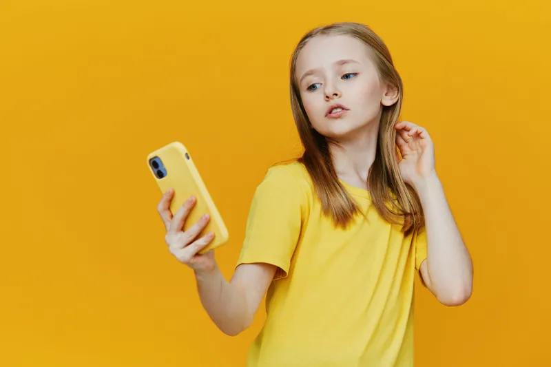 Young girl with long hair wearing a bright yellow shirt, holding a yellow phone against a vibrant yellow background, expressing a thoughtful emotion
