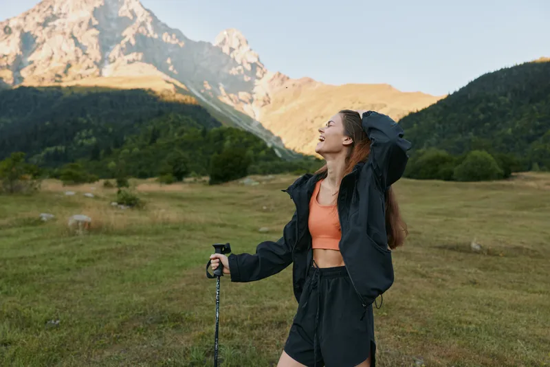 A cheerful hiker stands in a wide meadow with rugged mountains behind, looking up with a relaxed smile. Athletic jacket and shorts suit the outdoor scene.