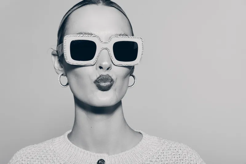 Stylish white woman with pale skin wearing large sunglasses and hoop earrings blows a kiss. Fashion portrait with confident mood in monochrome studio.