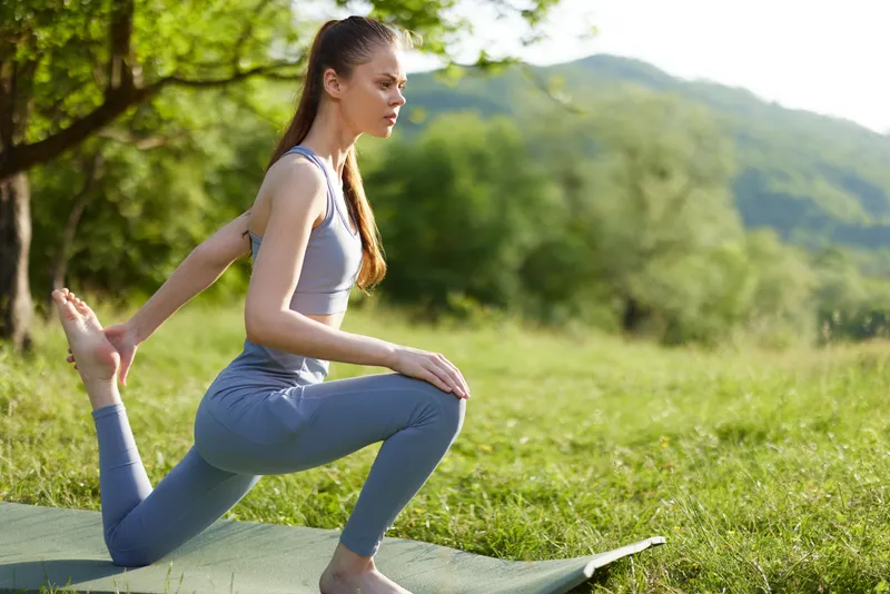 Young woman practicing yoga outdoors in nature, wearing light gray athletic clothing, showcasing a peaceful expression in a green landscape