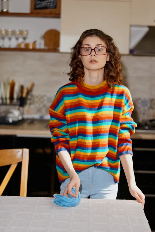 Woman with glasses in a rainbow striped sweater cleans a kitchen counter, wearing a blue glove, focused on tidying a bright home scene, casual lifestyle moment