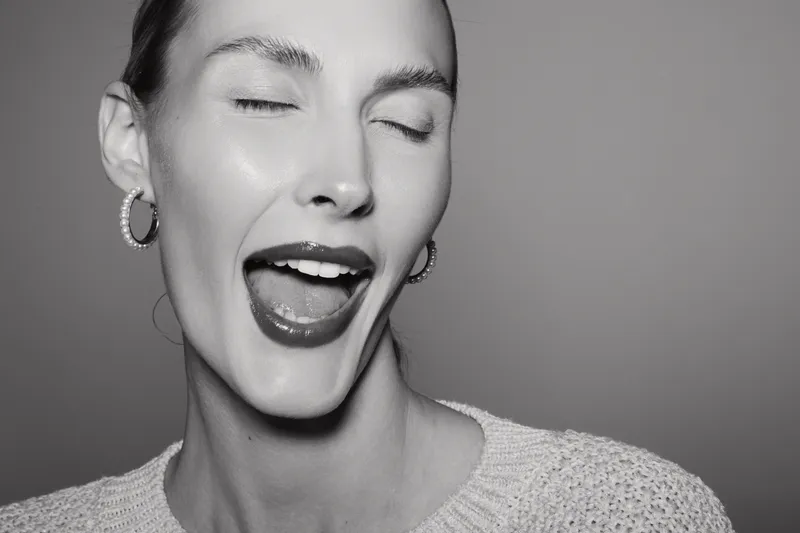 Close up of young caucasian woman with fair skin winking and smiling, wearing hoop earrings and sweater on grey background.