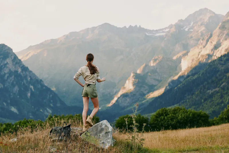 Woman with ponytail standing on rock in mountain field, looking at distant peaks. Outdoor lifestyle, adventure, nature exploration, freedom, and hiking concept.