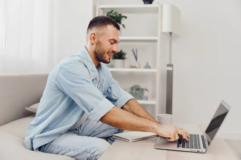 Smiling man working on laptop, enjoying a productive moment at home. Bright, cozy interior with plants fosters a relaxed atmosphere for creativity and focus.