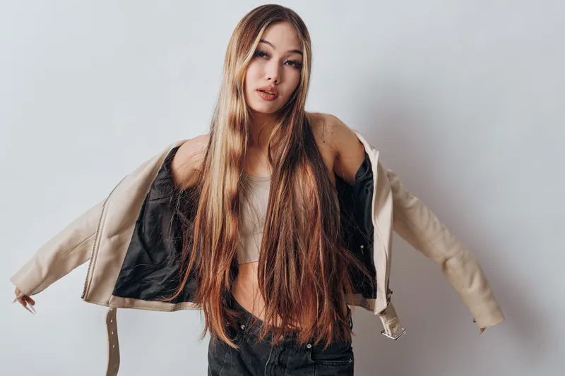 Young woman with long straight hair putting on beige jacket over white top in studio. Beauty, fashion and style with casual outfit and confident pose on white background.